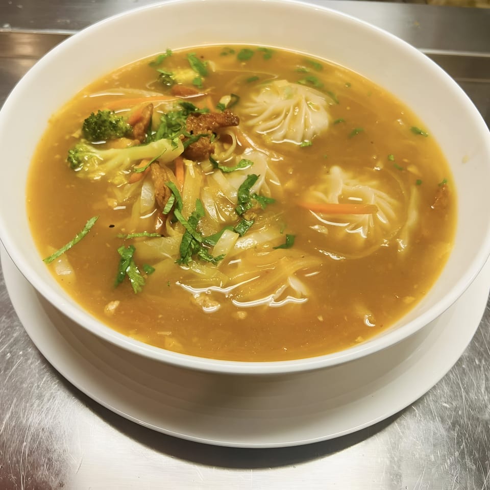 A bowl of Indian momo soup with vegetables, noodles, and dumplings in a rich broth.